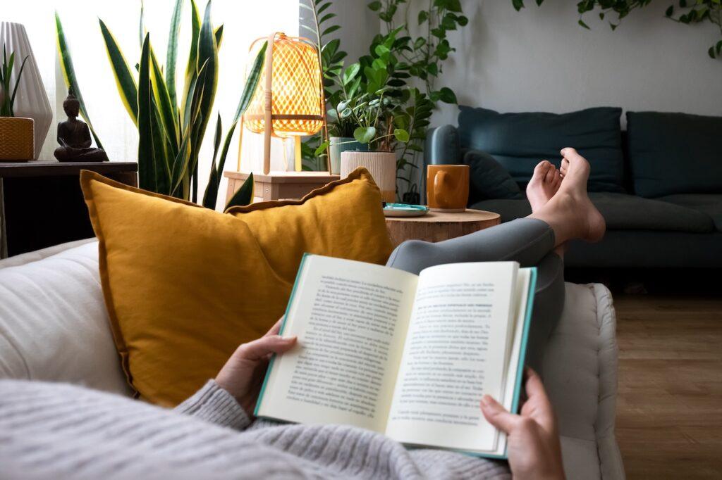 Person liest ein Buch auf einer gemütlichen Couch in einem Wohnzimmer mit vielen Pflanzen, gelben Kissen und warmem Ambiente